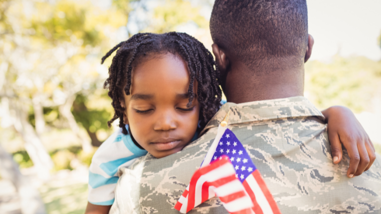 A Veteran in uniform embracing their child, who is holding a small American flag